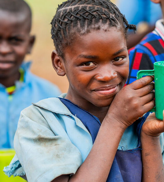Image of a child holding her mug up to the camera, with other children pictured in the background. 